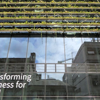 Glass-fronted building with reflections of the sky and nearby structures. Rows of greenery are integrated into the design above the windows. Large text on the building's facade reads 'Transforming Business for Good.'